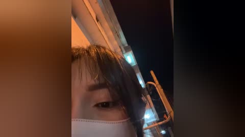 Media: Video of a young Asian woman with short brown hair, wearing a surgical mask, peering through a window with a blurred cityscape in the background.