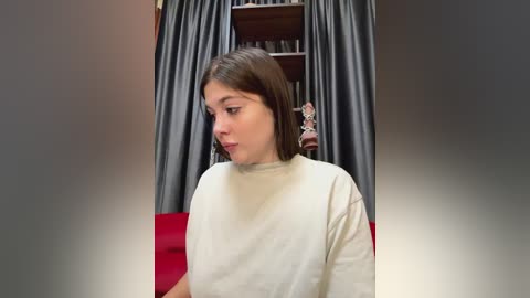 Media: Video of a young woman with shoulder-length brown hair, wearing a white sweater, standing in a room with grey curtains and a red couch.