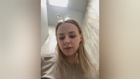 Media: A video of a young Caucasian woman with fair skin and long blonde hair, wearing a beige top, captured from a low angle in a narrow, beige-walled hallway with a ceiling light.