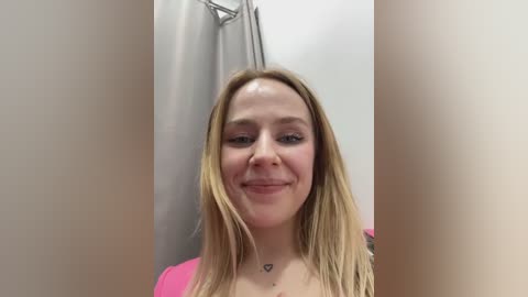 Media: A video of a smiling young Caucasian woman with straight blonde hair, light skin, and wearing a pink top, standing in a bathroom with a metal shower rod.