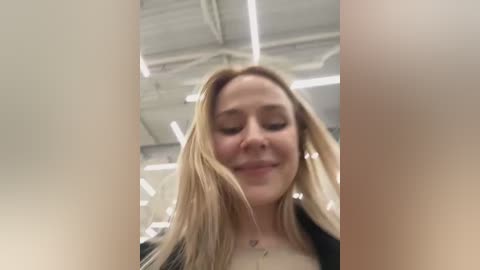 Media: A video of a blonde woman with light skin, wearing a black jacket, smiling slightly, taken indoors with a ceiling of exposed pipes and fluorescent lighting.