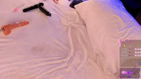 Media: Video of a white pillow with a beige sheet, featuring a black dildo, a pink vibrator, and an orange dildo on the bed, alongside a smartphone screen displaying a game interface.