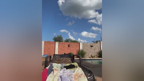 Media: A video of a sunlit outdoor patio with a pool, featuring a lounging area with a white and yellow blanket, surrounded by a red brick wall under a clear blue sky.