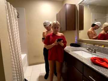 Media: Video of a modern bathroom with a blonde woman in a red dress and cap taking a selfie, a shirtless man in a cap looking at her in the background.