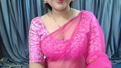 Media: Video of a woman with light skin, wearing a vibrant pink sari with intricate floral embroidery, against a backdrop of gray, draped fabric. She has a small, delicate gold necklace and her dark hair is partially visible.