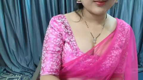 Media: Video of an Indian woman with fair skin, wearing a pink saree with white floral designs, and a gold necklace, against a gray curtain background.