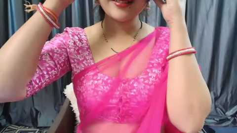 Media: Video of a light-skinned woman in a bright pink saree, adorned with intricate floral embroidery, wearing gold bangles and a thin necklace. Background features gray curtains.