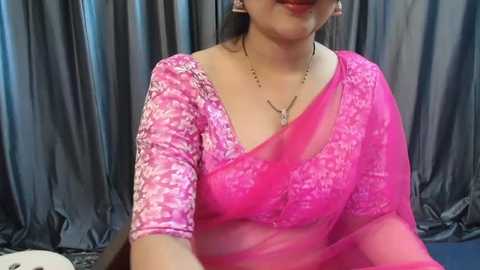 Media: A video of a South Asian woman with medium skin tone, wearing a bright pink saree with a floral design, seated against a backdrop of gray, flowing curtains. She has long, dark hair and wears a gold necklace.