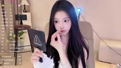 Media: A video shows a young Asian woman with long black hair, fair skin, and makeup, holding a black phone with a safety symbol. She sits in a beige chair, in a room with a white lamp and a blue light.