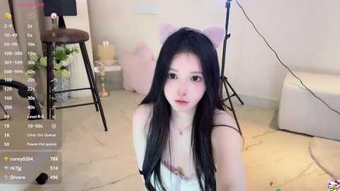 Media: Video of a young East Asian woman with long black hair and pale skin, wearing a white dress and pink cat ears, sitting on a beige carpet in a modern living room.