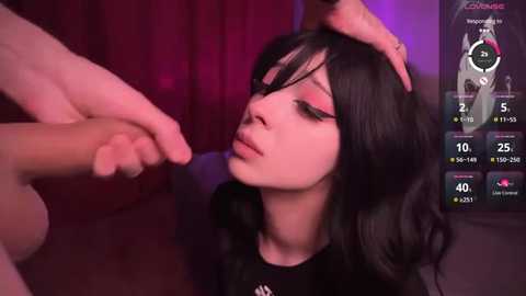 Media: Video of a young Asian woman with black hair and pink eyeshadow, wearing a black top, being caressed by a hand, in a dimly lit room with a purple light.