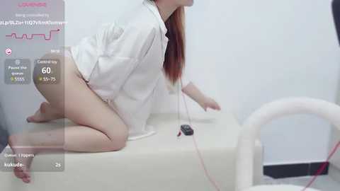 Media: Video of a light-skinned woman with long red hair, wearing a white robe, kneeling on a white cushion in a white room. A black microphone and red cable are nearby.