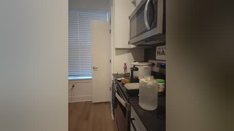 Media: Video of a narrow, compact kitchen with white cabinets, a microwave, and a stove, partially visible through an open doorway. The room has hardwood flooring and a small window with white blinds.