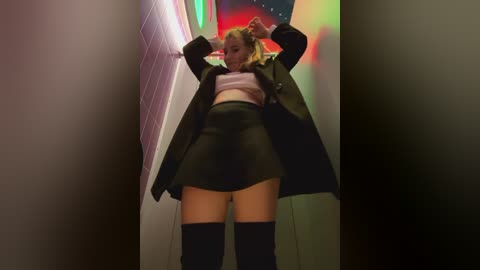 Media: Video of a young woman with blonde hair, wearing a black coat, white crop top, black skirt, and thigh-high boots, posing in a brightly lit, narrow hallway with colorful neon lights and a tiled wall.