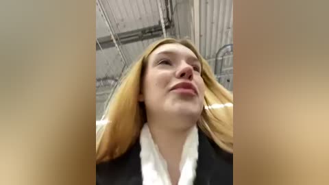 Media: Video of a young, fair-skinned woman with long, straight blonde hair, wearing a black blazer over a white shirt, looking up with a neutral expression. Background shows a metal ceiling and industrial lighting.