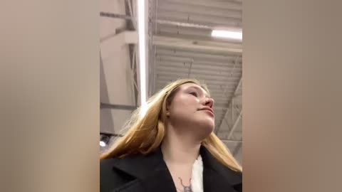 Media: A video of a young woman with long blonde hair, fair skin, and wearing a black blazer, standing in an industrial, dimly-lit room with exposed beams and pipes.