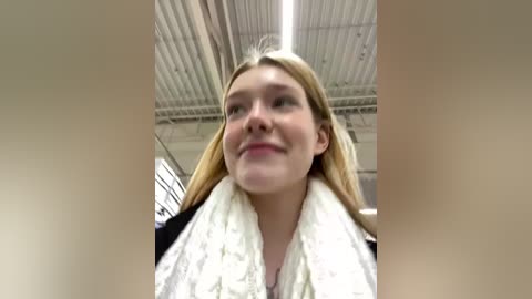 Media: Video of a young Caucasian woman with long blonde hair, wearing a white towel, standing in a large industrial warehouse with a corrugated metal ceiling.