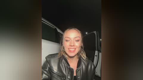 Media: Video of a smiling, blonde, fair-skinned woman with shoulder-length hair, wearing a black leather jacket, sitting in a dark, open SUV at night.