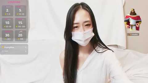 Media: Video of an Asian woman with long black hair, wearing a white mask and light-colored top, in a hospital-like setting.