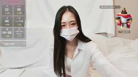 Media: A video of an Asian woman with long black hair, wearing a white surgical mask and a light blue shirt, posing in a white bed.