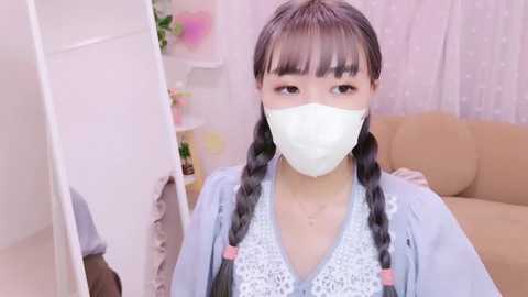 Media: Video of an Asian woman with long braided hair, wearing a light blue crocheted top, white face mask, and a light blue jacket, indoors with pink and white decor.