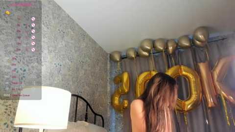 Media: A video of a woman with long brown hair, partially visible from behind, in a room with gold \"2016\" balloons, a white lamp, and a tiled wall.