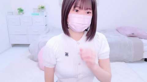 Media: A video of a young East Asian woman with shoulder-length dark hair and fair skin, wearing a white mask, white blouse, and a pink shirt. She stands in a minimalist white bedroom with a bed, dresser, and potted plants in the background.