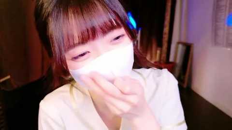 Media: Video of an Asian woman with straight, dark hair and bangs wearing a white mask, holding her nose, looking distressed, indoors with dim lighting.