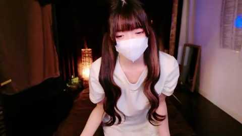 Media: Video of an Asian woman with long, wavy brown hair, wearing a white mask, a white shirt, and a white skirt, sitting indoors with dim lighting and a lit lantern in the background.