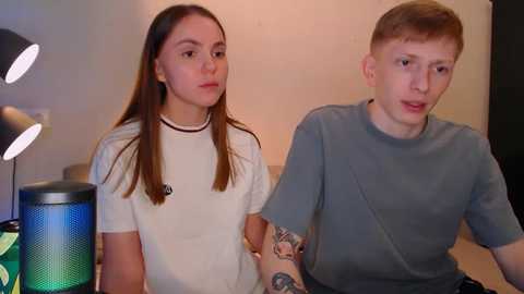 Media: Video of a young Caucasian couple, a girl with long brown hair and a boy with short red hair, wearing casual clothes, seated in a dimly lit room with a green lamp and a beige wall.