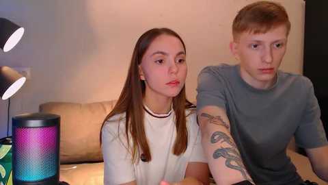 Media: Video of two young white people with straight brown hair, one wearing a black choker, sitting closely on a beige couch, illuminated by a colorful lamp and a dark lampshade.