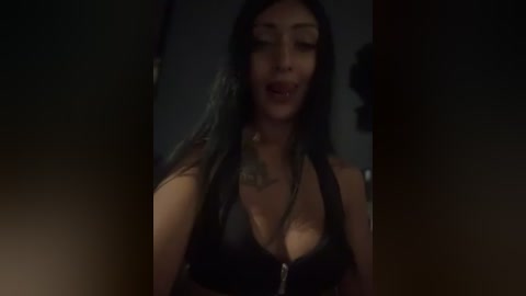 Media: Video of a woman with long, black hair in a dimly lit room, wearing a black, plunging neckline top, and a tattoo on her neck.