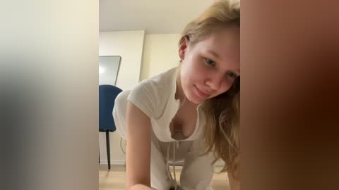 Media: A video of a young, slender, Caucasian woman with long blonde hair, wearing a white top, bending over, peering through a gap between two large, brown objects, in a minimalist room with a blue chair and a mirror.