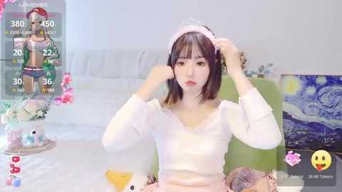 Media: Video of a young Asian woman with short brown hair, wearing a white crop top, adjusting a pink headband in a modern, minimalistic room. Virtual overlays show gaming stats and emojis.