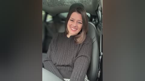 Media: Video of a smiling woman with shoulder-length brown hair, wearing a dark knit sweater, sitting in a car with black leather seats and a gray headrest.