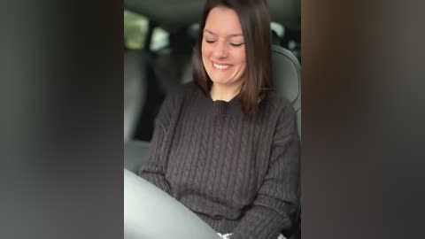 Media: Video of a young Caucasian woman with straight, dark brown hair, wearing a dark grey, textured sweater, smiling while driving a car with grey seats.