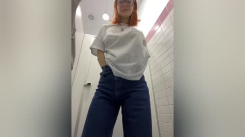 Media: Video of a young woman with shoulder-length red hair, wearing a loose white t-shirt and high-waisted blue jeans, standing in a narrow, tiled bathroom with white walls and a metal door.