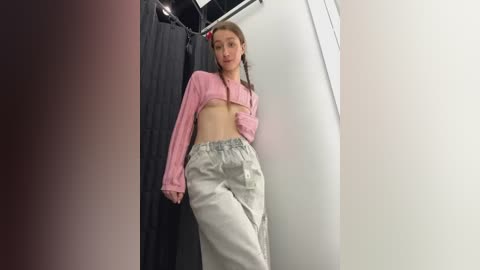 Media: A video of a young woman with fair skin and straight brown hair, wearing a pink crop top and high-waisted beige pants, posing in a fitting room with a black curtain and white wall.