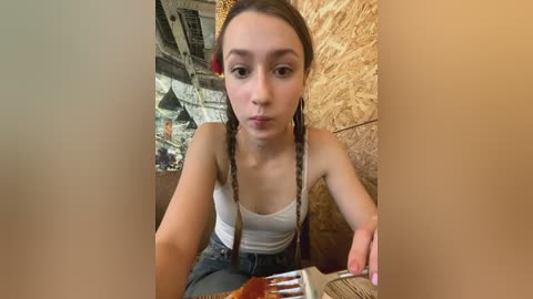 Media: Video of a young, fair-skinned woman with long, brown pigtails, wearing a white tank top and jeans, eating a burger. Background features a wooden wall and a dimly lit, industrial-themed room with metal structures.