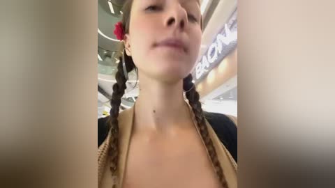 Media: A video of a fair-skinned woman with light brown hair styled in two braids, wearing a black top, looking down at a cellphone screen. The background shows a blurred airport setting with a \"BLOW\" sign.