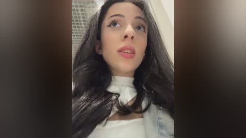 Media: Video of a young woman with long, dark hair, wearing a white top, looking upward with a neutral expression. Background features a tiled wall and blurred brown objects.