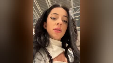 Media: Video of a young woman with long, straight black hair, wearing a white turtleneck sweater, looking straight into the camera with a neutral expression. The background shows an industrial ceiling with exposed beams.