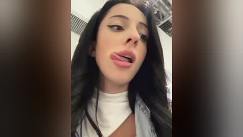 Media: Video of a young woman with long black hair, wearing a white top and denim jacket, sticking her tongue out playfully. Background shows a ceiling with vents.