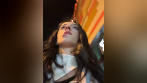 Media: Video of a woman with long brown hair, wearing a white dress, looking upward, with a blurred background of a colorful, possibly striped structure.