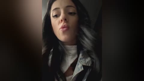 Media: Video of a young woman with long black hair, fair skin, and a neutral expression. She wears a white turtleneck under a denim jacket, set against a dark background.