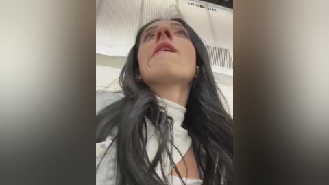 Media: Video of a woman with long black hair and pale skin, wearing a white top, looking upwards with a slight smile. Background features a ceiling with a vent and lighting.