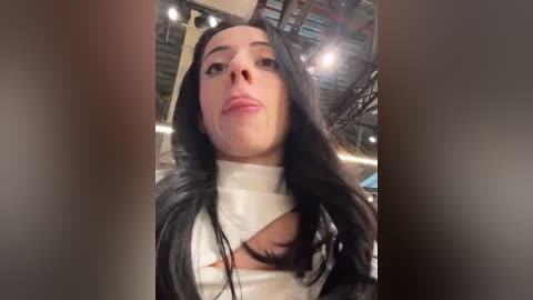 Media: Video of a young woman with long, straight black hair, wearing a white turtleneck with a large, partially cut-out design, revealing a glimpse of her chest. Background features industrial elements, possibly a warehouse.