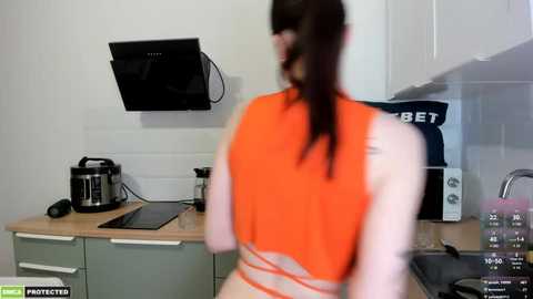 Media: A blurry video of a person in an orange tank top with long hair, standing in a modern kitchen with appliances, white cabinets, and a \"BET\" sign.