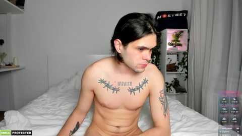 Media: Video of a shirtless, pale-skinned man with long black hair and tattoos, sitting on a white bed in a simple bedroom.