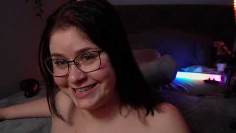 Media: Video of a smiling, fair-skinned woman with dark hair, wearing glasses, topless, lying on a bed with grey sheets, in a dimly lit room with neon lights in the background.